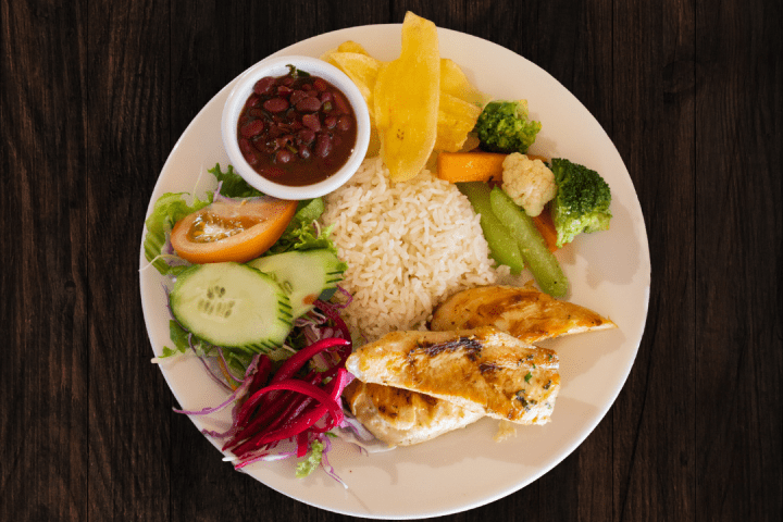 Plate with rice, grilled chicken, vegetables, plantains, beans, and salad on a wooden table.