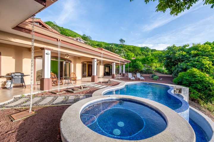 Luxury house with patio, pool, and greenery under blue sky.