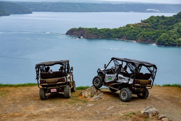 Two off-road vehicles parked on a cliff overlooking a scenic bay with lush greenery.