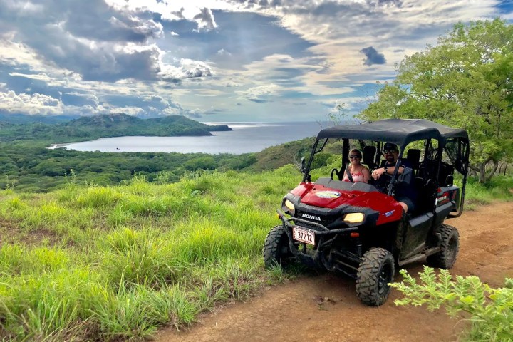 ATV with two people on a dirt path, overlooking a scenic hilly landscape and ocean view.