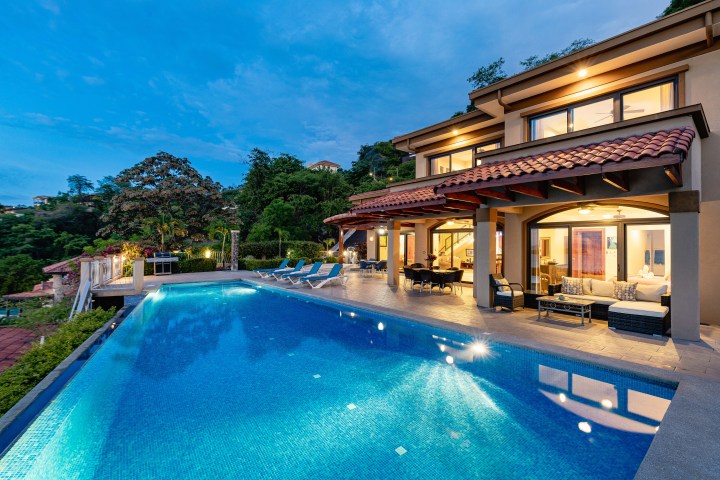 Luxurious villa with infinity pool, patio furniture, and garden, set against a twilight sky.