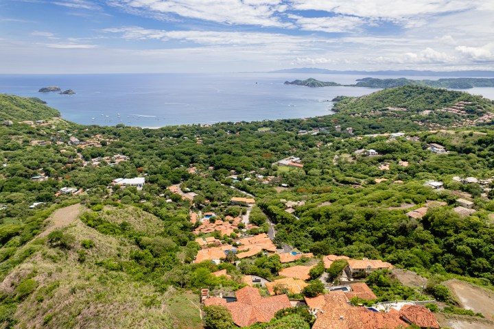 Aerial view of a coastal town with lush greenery and ocean in the background under a partly cloudy sky.