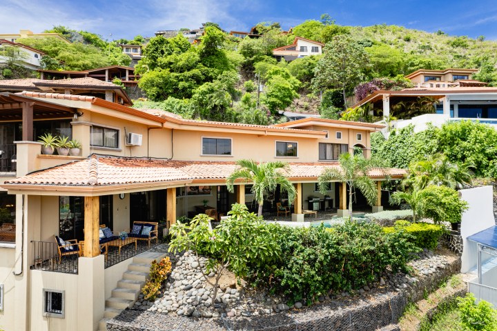House with terracotta roof, patio, and tropical plants on a hillside.