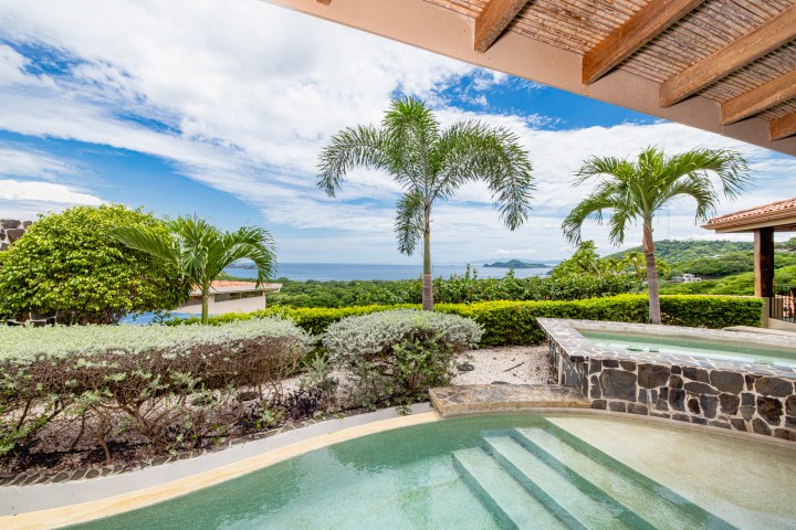 Tropical patio view with a pool, palm trees, and ocean under a partly cloudy sky.
