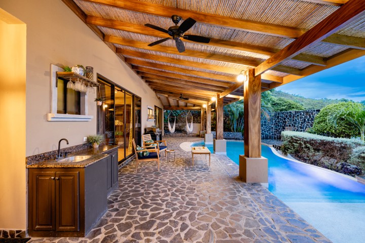 Covered outdoor patio with stone flooring, lounge chairs, a sink, and adjacent pool in a tropical setting.