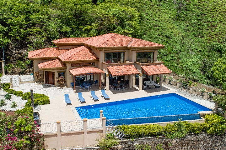 Large house with orange roof, pool, surrounded by greenery and patio furniture in garden.