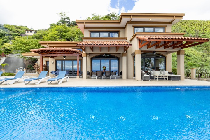 Luxury villa with pool, patio furniture, and sun loungers, surrounded by greenery.