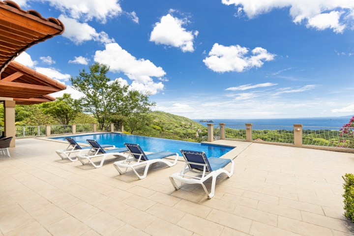 Poolside view with lounge chairs, overlooking ocean and greenery under a blue sky.