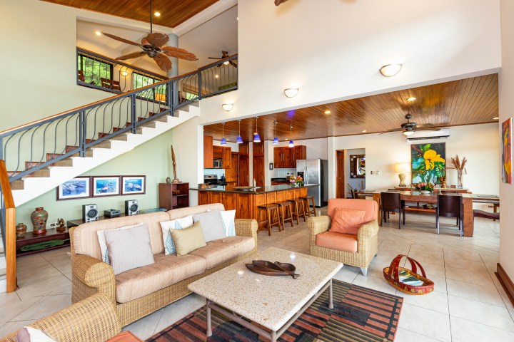 Spacious living room with wicker furniture, dining area, and staircase, featuring wooden ceiling and bright decor.