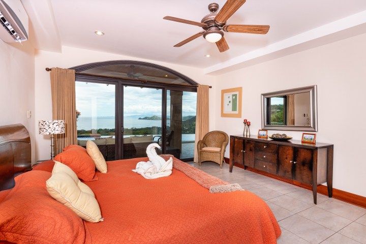 Bedroom with orange bedding, ceiling fan, and ocean view through large glass doors.