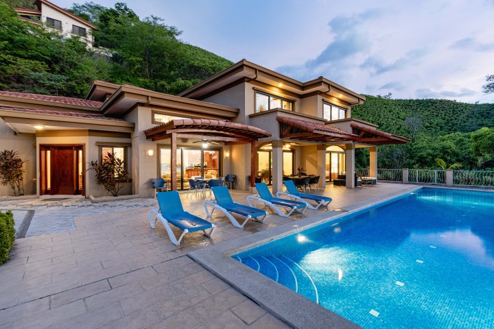 Luxury villa with pool, patio, and sun loungers at dusk in a lush hillside setting.