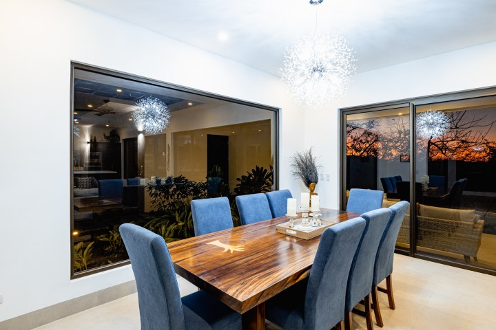 Dining room with blue chairs and wooden table, lit by spherical chandeliers, window view of sunset.