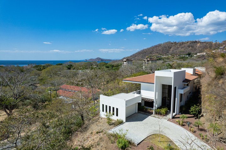 Modern white house with red-tiled roof on a hillside overlooking the ocean and distant mountains.
