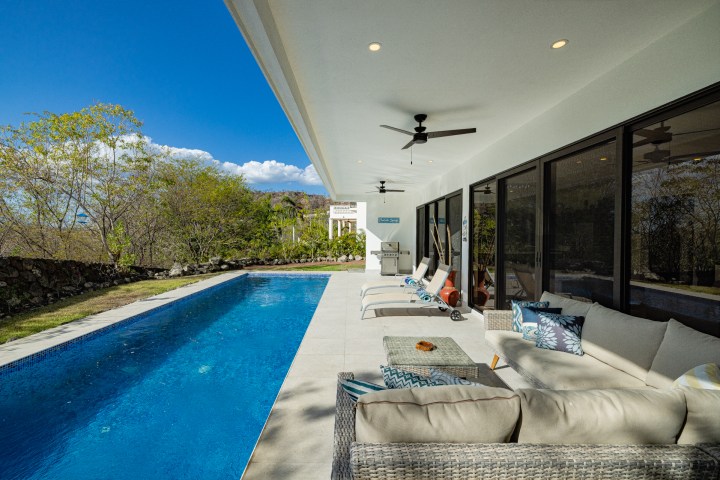 Modern patio with pool, lounge chairs, and sofa under a clear blue sky.