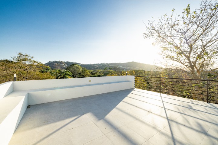 Sunny rooftop terrace with railings, trees, and distant hills.