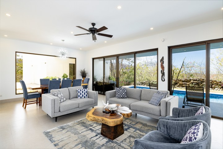 Modern living room with gray furniture, wooden table, and large windows overlooking a pool and garden.
