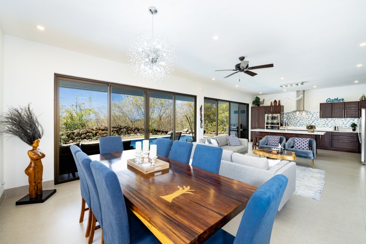 Open-plan living area with dining table, blue chairs, couches, kitchen, and large windows.