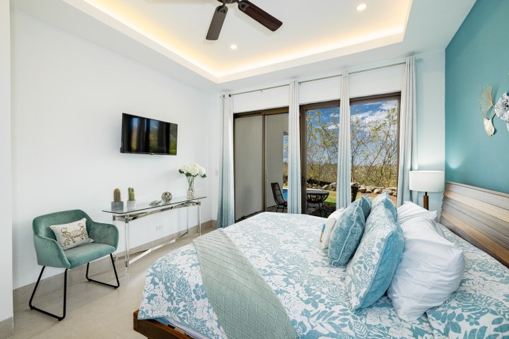 Bright bedroom with blue accents, large window, and modern decor, featuring a bed, TV, and chair.