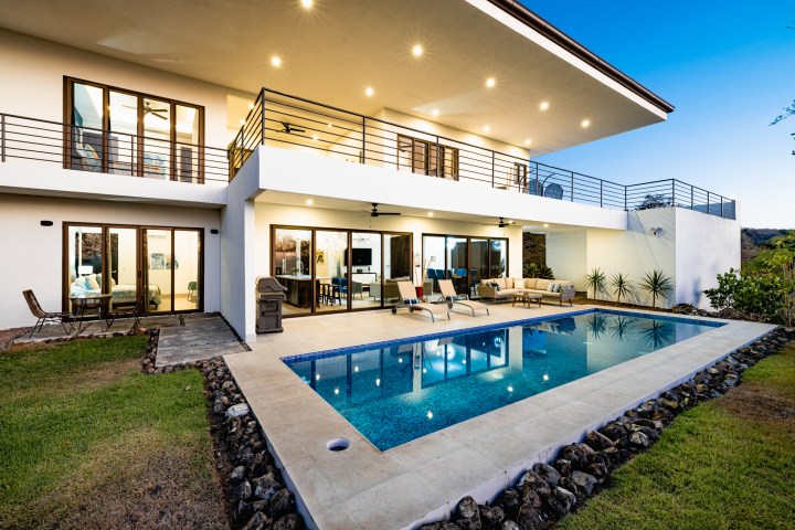 Modern two-story house with pool, patio, and balcony at dusk.