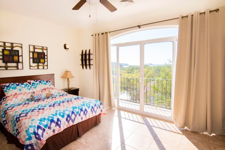 Bright bedroom with patterned bedspread, art on walls, and large window overlooking trees.