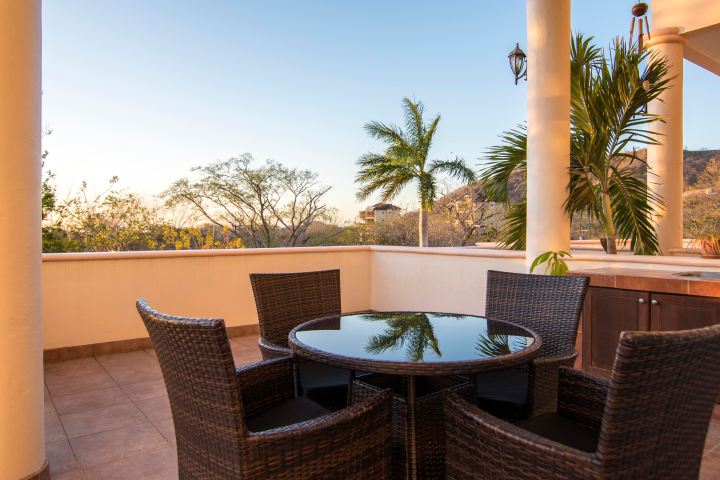 Outdoor patio with wicker table and chairs, palm trees, and mountain view.