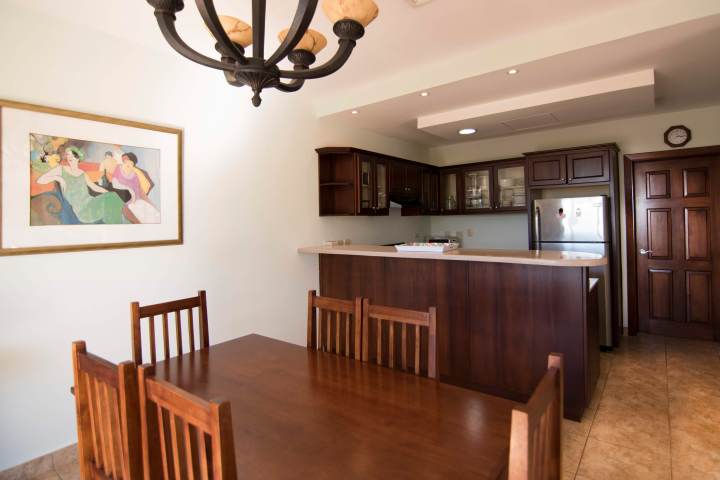 Dining room with wooden table, chairs, kitchen with cabinets, artwork, and chandelier.