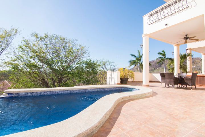 Outdoor pool on a sunny terrace with trees and a dining area under a covered patio.
