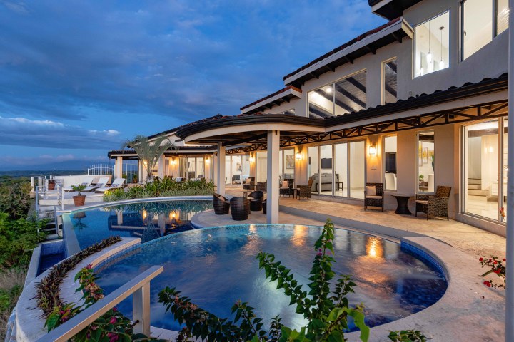 Luxury home exterior with infinity pool and patio chairs at sunset.