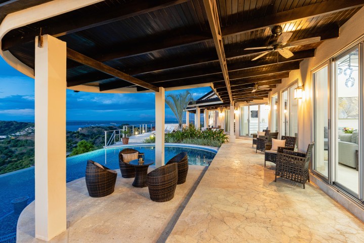 Luxurious patio with poolside seating, ocean view, ceiling fans, and warm lighting at dusk.