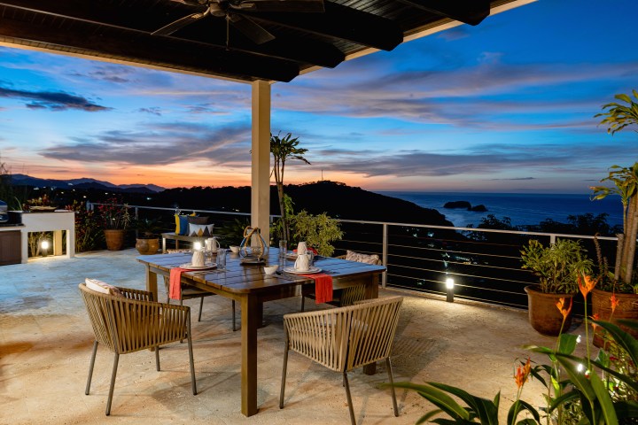 Outdoor dining area overlooking ocean sunset with table, chairs, and plants.