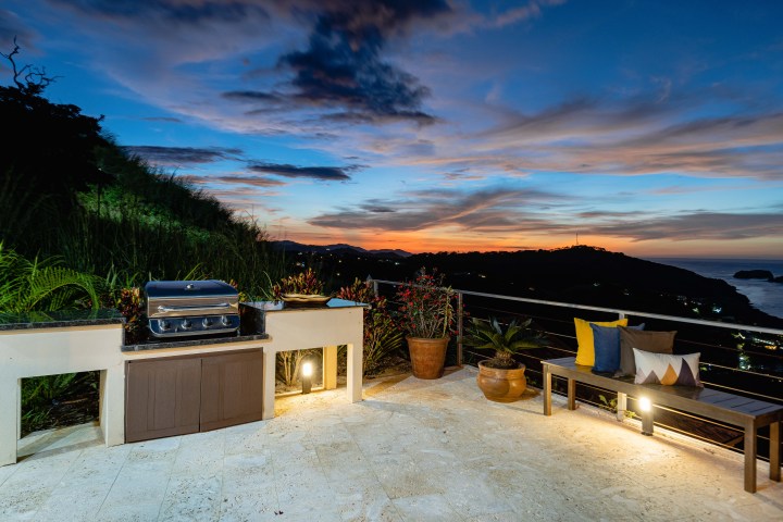 Outdoor patio with grill and seating at sunset, overlooking hills and ocean.