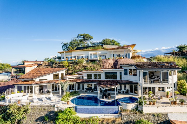 Modern hillside villa with pool, large windows, and terracotta roof tiles, under a clear blue sky.
