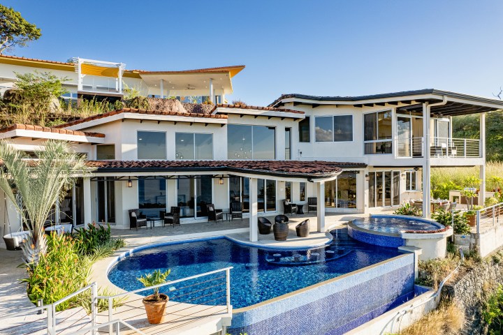 Modern hillside house with blue pool, large windows, and lush greenery under a clear sky.