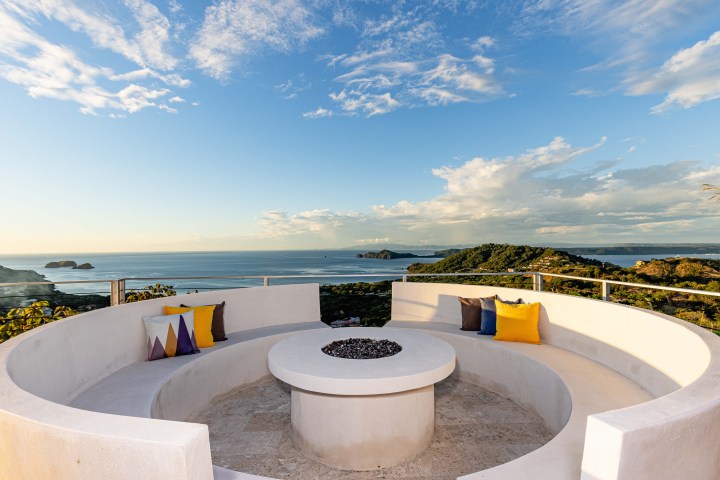 Curved outdoor seating with colorful pillows overlooks ocean and islands under a partly cloudy sky.
