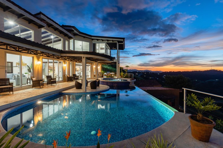 Luxury house with large pool at sunset, showcasing lit windows and panoramic views.