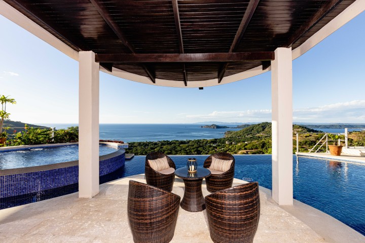 Covered patio with wicker chairs and ocean view, overlooking infinity pool and hillside.