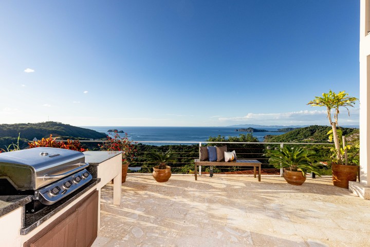 Outdoor patio with grill, bench, potted plants, and ocean view under a clear blue sky.