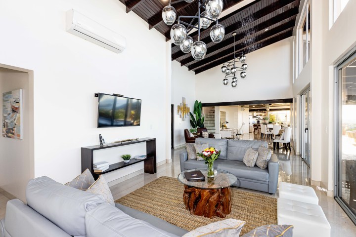 Modern living room with gray sofas, glass table, TV, and chandelier, open to dining area with large windows.