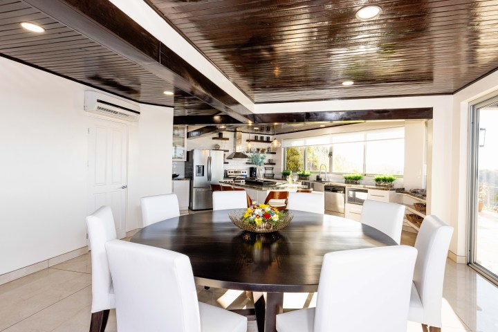 Modern kitchen with round table, white chairs, dark wood ceiling, and stainless steel appliances.
