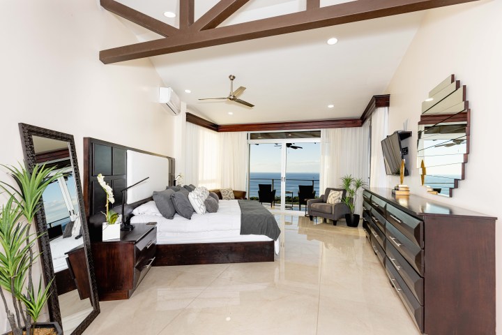 Modern bedroom with ocean view, large bed, mirrors, dresser, and plants.