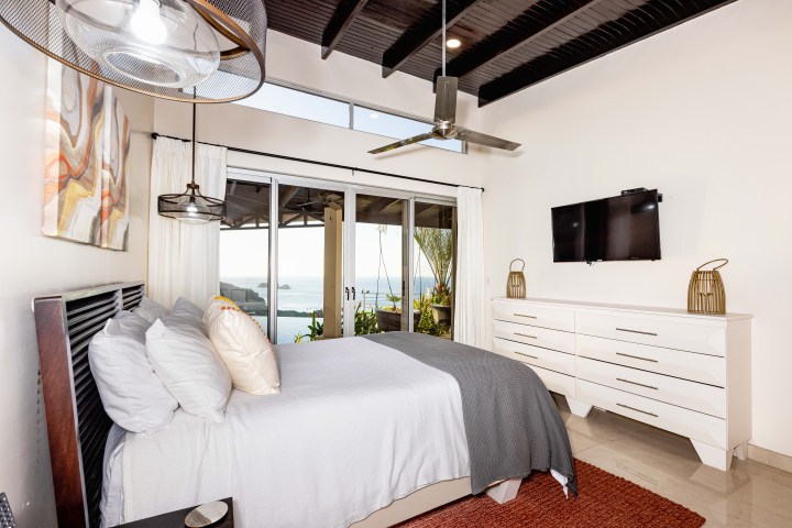 Modern bedroom with ocean view, featuring a bed, dresser, TV, artwork, and ceiling fan.