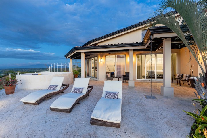 Luxury patio with three lounge chairs, overlooking scenic view and modern house at dusk.
