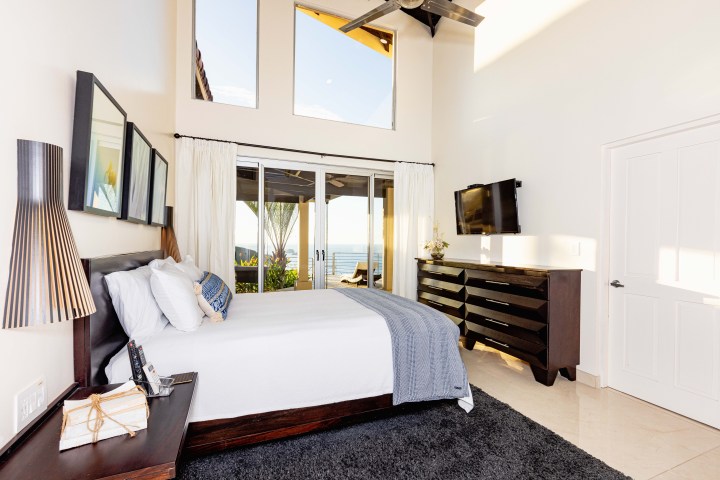 Bright modern bedroom with a bed, dresser, wall art, and large windows overlooking a balcony and sea view.