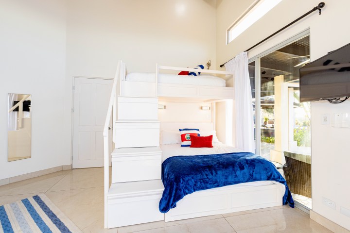 Bright room with white bunk bed, blue bedding, and a wall-mounted TV beside a glass door.