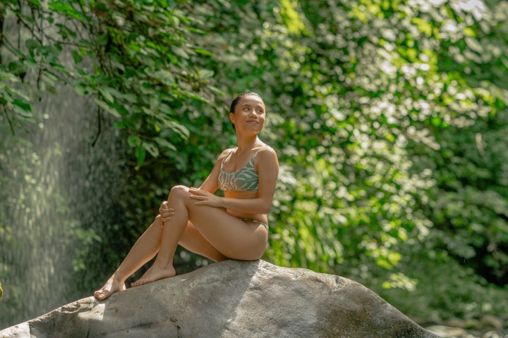 Woman in swimsuit sitting on rock near waterfall and lush greenery.