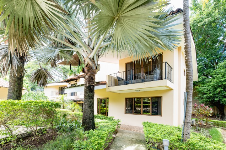 Tropical garden view of a beige two-story house with large windows and balcony.