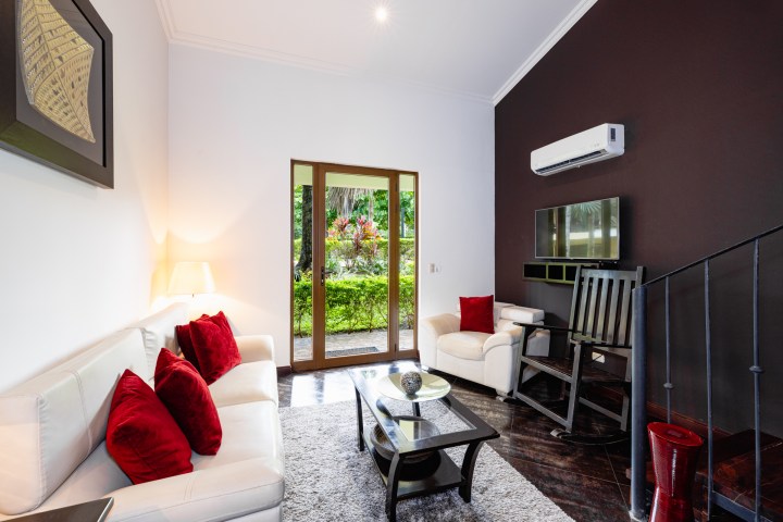 Modern living room with white sofas, red pillows, glass doors, and an air conditioner on a dark accent wall.