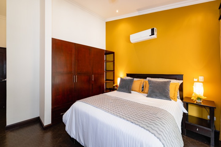 Modern bedroom with mustard accent wall, bed with gray and mustard pillows, wooden wardrobe, and air conditioner.