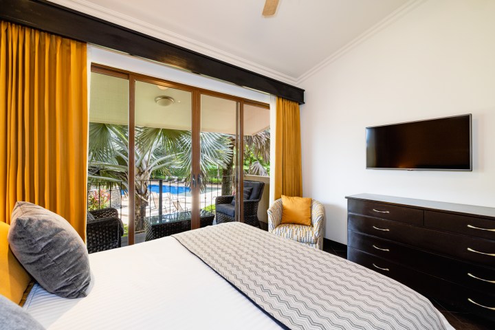 Bedroom with TV, dresser, yellow curtains, palm view, and pool outside sliding door.