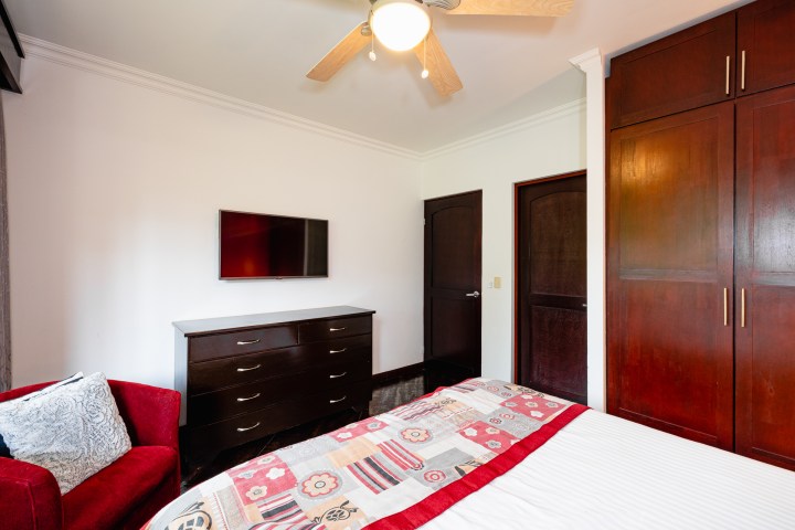 Bedroom with red sofa, patterned bedspread, TV on dresser, ceiling fan, and dark wood furniture.